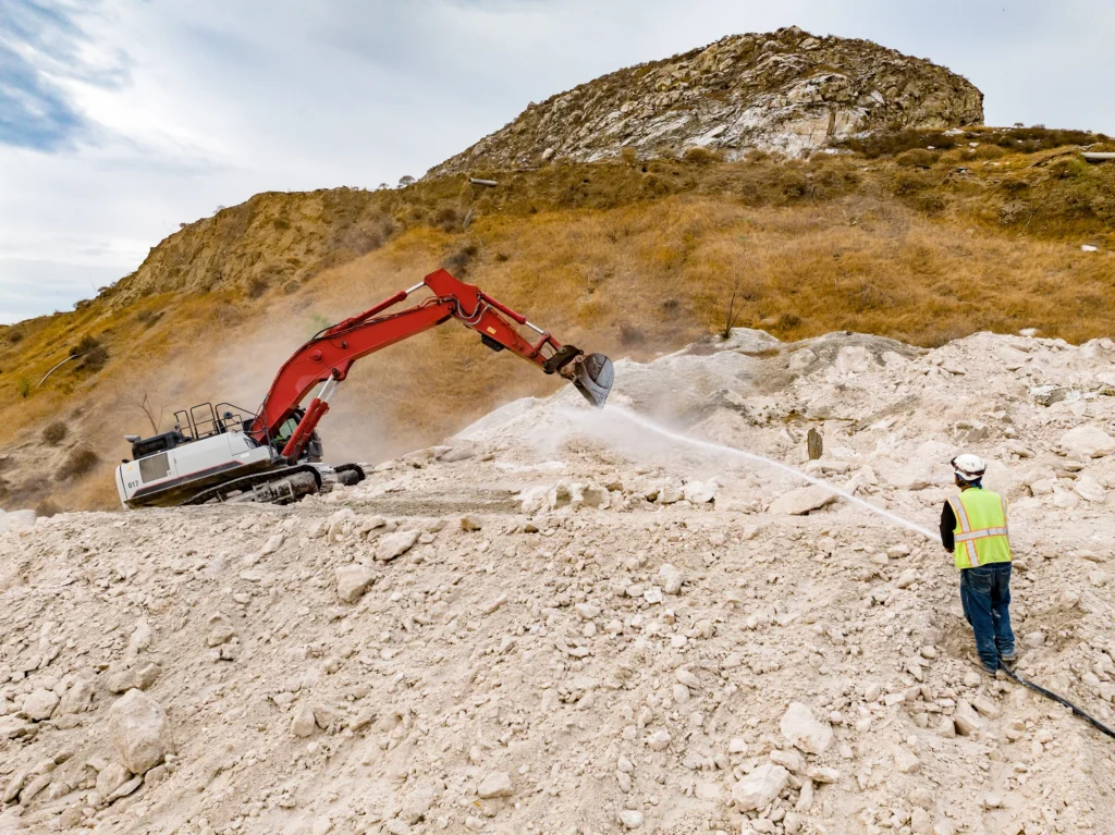 Water being sprayed to suppress dust while an excavator breaks rock.