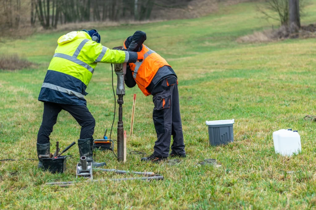 Two workers using portable soil sampling equipment on a grassy site.